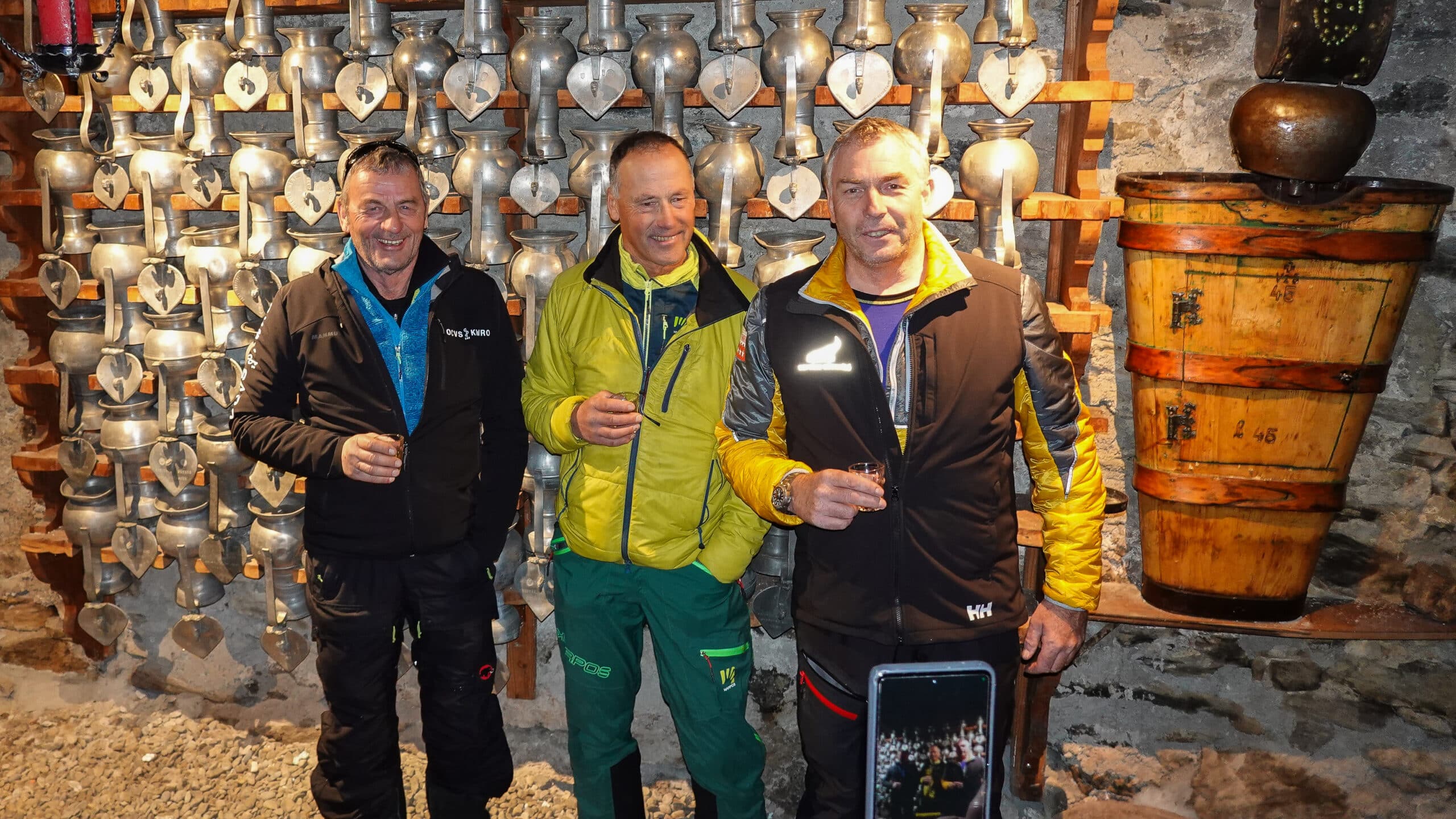 Trois hommes souriants devant cloches traditionnelles et un tonneau.