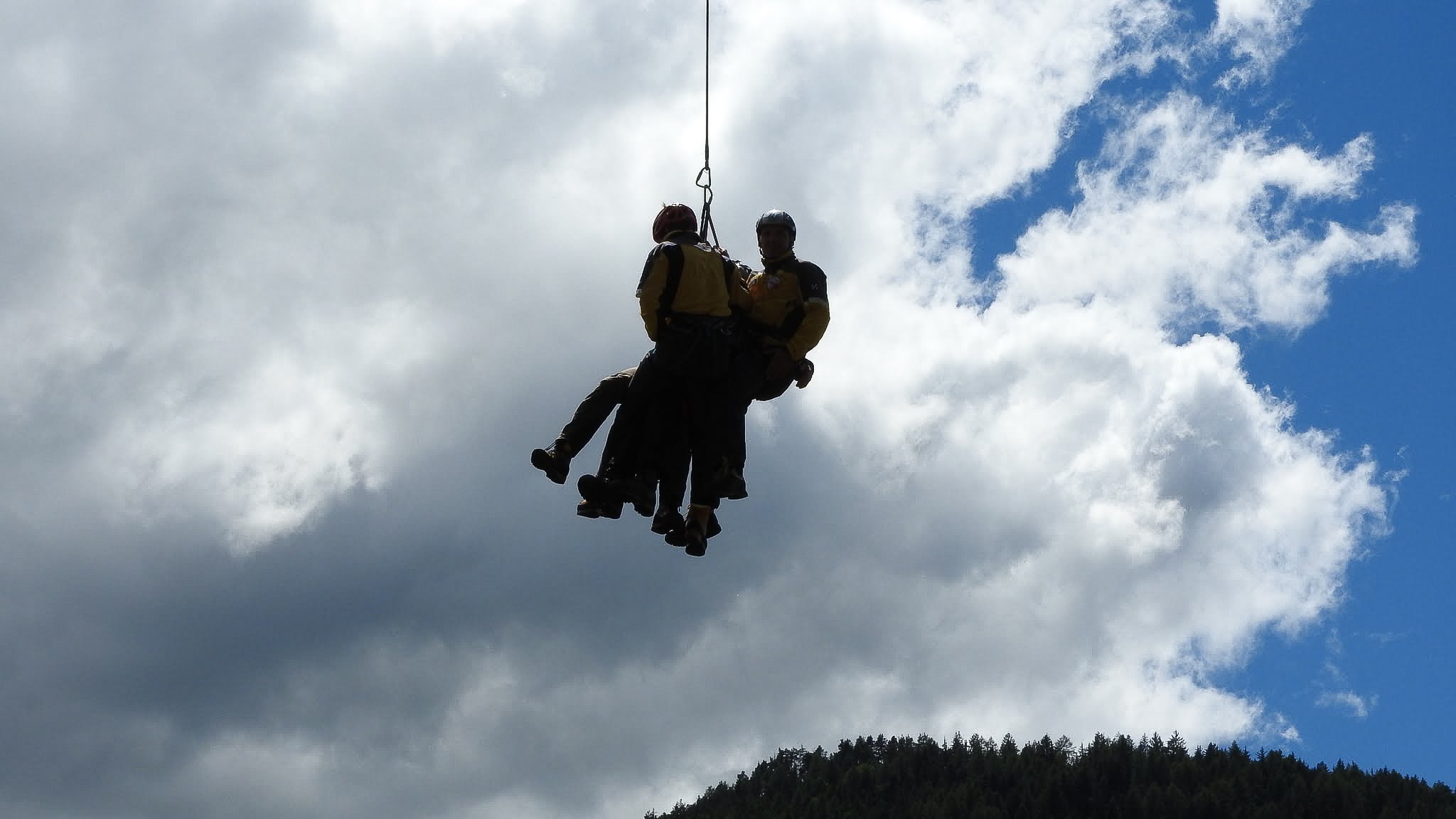 Deux secouristes héliportent une personne au-dessus d'une forêt montagneuse.