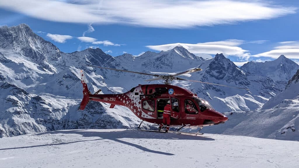 Hélicoptère rouge Air Zermatt sur neige, montagnes enneigées majestueuses.