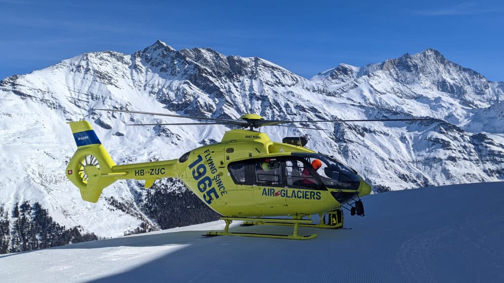 Hélicoptère jaune Air Glaciers posé sur neige, montagnes enneigées en arrière-plan.
