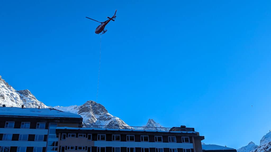 Hélicoptère transportant une charge au-dessus d'un bâtiment et montagnes enneigées.